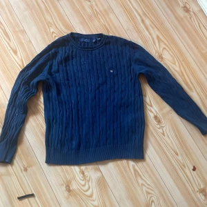 Mörkblå kabelstickad tröja från Gant - Säljer en mörkblå kabelstickad tröja från Gant med rund halsringning och diskret logga på bröstet. Tröjan har långa ärmar och ribbade muddar vid ärmslut och nederkant. Perfekt för dig som gillar klassisk stil.