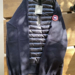Marinblå HyBridge Knit Packable Pullover från Canada Goose - Säljer en stilren marinblå HyBridge Knit Packable Pullover från Canada Goose. Jackan har en quiltad framsida med dragkedja och ett broderat märke på ärmen. Perfekt för kyliga dagar med sin kombination av stil och funktionalitet.
