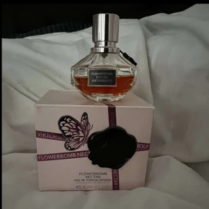 Flowerbomb Nectar från Viktor & Rolf - Säljer en exklusiv parfym, Flowerbomb Nectar från Viktor & Rolf. Flaskan har en elegant, facetterad design med en silverfärgad kork. Doften är intensiv och blommig, perfekt för den som vill sticka ut. Förpackningen är ljusrosa med en dekorativ fjäril och ett svart sigill.
