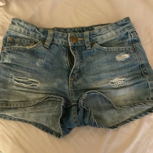 Blå jeansshorts från Sunny Star - Säljer ett par coola blå jeansshorts från Sunny Star med slitna detaljer och klassisk femficksdesign. Perfekta för en avslappnad stil. De har en knapp och dragkedja framtill.