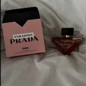 Paradoxe Eau de Parfum från Prada - Säljer en elegant Paradoxe Eau de Parfum från Prada. Flaskan har en unik triangulär form i en varm bärnstenston med en svart kork. Förpackningen är stilren i rosa med svart text. Perfekt för den som vill ha en lyxig doftupplevelse.