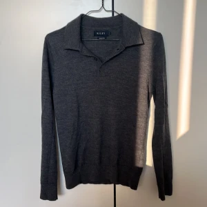 Grå merinoull polotröja från Riley - Mörkgrå långärmad polotröja från Riley i mjuk merinoull. Klassisk krage med knäppning framtill. Den är knappt använd så skicket är fortfarande mycket bra. Storleken är s men den är liten i storleken så jag skulle mer säga att den är en xs. 
