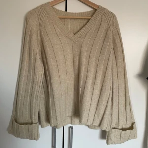 Beige ribbstickad tröja med v-ringning - Mysig beige stickad tröja med v-ringad hals och ribbat mönster. Tröjan har långa ärmar med uppvikta muddar och en avslappnad passform.
