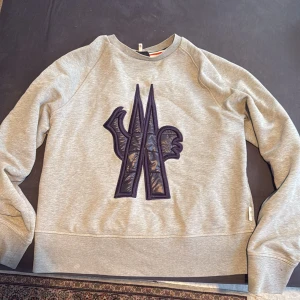Grå sweatshirt från Moncler med stor logga - Säljer en grå sweatshirt från Moncler med en stor, vadderad svart logga framtill med likadant material som en jacka. Tröjan har rund halsringning och långa ärmar. Perfekt för dig som gillar exklusiva streetwear-plagg.