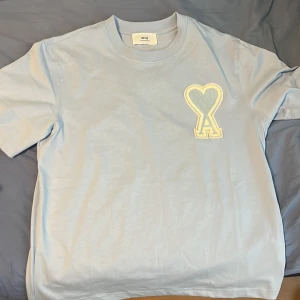 Ljusblå t-shirt från Ami Alexandre Mattiussi - Snygg ljusblå t-shirt från Ami Alexandre Mattiussi med ett stort broderat hjärta och A-logga på bröstet. Klassisk passform och rund halsringning. Tillverkad i mjuk bomull för extra komfort.