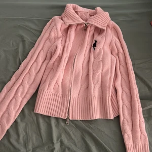 Rosa stickad kofta från SHEIN - Säljer en söt rosa stickad kofta från SHEIN med dragkedja framtill och ribbade detaljer vid ärmslut och nederkant. Koftan har ett klassiskt flätmönster och en liten krage. Perfekt för kyliga dagar!