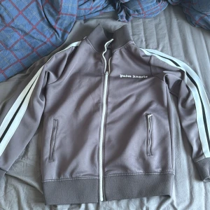 Kofta från Palm Angels - Säljer en kofta  från Palm Angels med vita ränder längs ärmarna och dragkedja framtill. Jackan har en hög krage och två fickor med dragkedja. Perfekt för en sportig look!