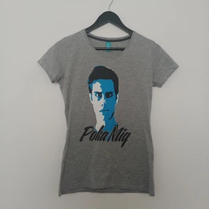 Grå t-shirt med tryck från IJWTBC - Säljer en grå t-shirt från IJWTBC med ett stort tryck av Victor's ansikte i blått och svart samt texten 'Poka Mig' på framsidan. T-shirten har rund hals och korta ärmar. Perfekt för dig som gillar IJustWantToBeCool