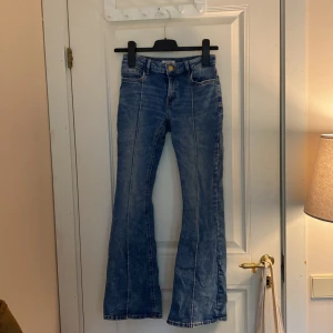 Blå bootcut jeans från Lindex - Säljer ett par klassiska blå jeans från Lindex med bootcut-passform. Jeansen har normal midja, fram- och bakfickor samt tydliga sömmar längs benen. 