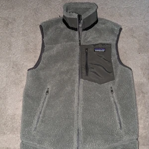 Grå fleeceväst från Patagonia - Säljer en grå fleeceväst från Patagonia med hög krage och dragkedja framtill. Västen har en bröstficka med dragkedja och två sidofickor. Perfekt för lager-på-lager och outdoor. Den passar om man är runt 160-165 cm lång!