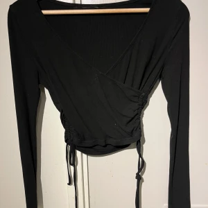 Svart omlottopp med knyt - Säljer en svart omlottopp med långa ärmar och knyt i sidan. Toppen har en v-ringad omlottdesign och är croppad, vilket ger en trendig look. Perfekt att styla med höga jeans eller kjol.