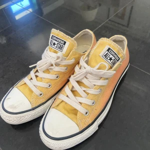 Gula och orange Converse All Star sneakers - Säljer ett par Converse All Star sneakers i tyg med snygg färgskiftning från gult till orange. Skorna har vita snören, vit tå och sula samt klassisk logga på plösen. Perfekta för dig som vill sticka ut med färg på fötterna!