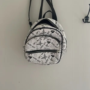 Vit ryggsäck med svart text och motiv - Säljer en vit ryggsäck med svarta axelremmar och tryck med text som 'baby girl', 'AMOR', 'BROKE' och olika motiv som hjärtan, fjärilar och ränder. Väskan har tre fack med dragkedja och är i fint skick.