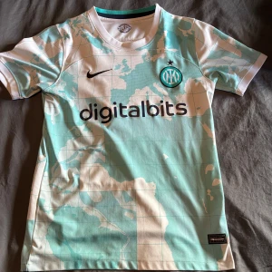 Inter Milan bortatröja 2022/23 - Snygg Inter Milan bortatröja från säsongen 2022/23 med digitalbits och Lenovo som sponsorer. Tröjan har ett unikt kartmönster i vitt och ljusblått, rund hals och svart Nike-logga. Perfekt för fotbollsfans som vill sticka ut.