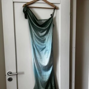 Grönblå långklänning i satin med knytband - Säljer en elegant långklänning i skimrande satin med vacker grönblå ombre-effekt. Klänningen har smala axelband och ett knytband på ena axeln för en unik detalj. Perfekt för speciella tillfällen!