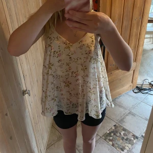Blommig v-ringad linnetopp - Söt och luftig linnetopp med blommigt mönster i ljusa färger. Toppen har v-ringning både fram och bak samt ärmlös design. Perfekt för varma dagar och ger en romantisk känsla.