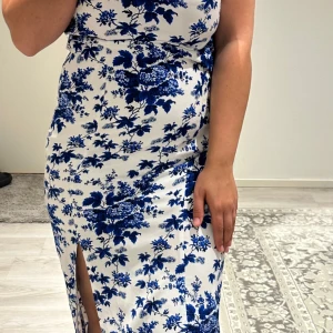 Vit och blå blommig klänning med slits - Säljer en vit klänning med blått blommönster och en snygg slits framtill. Klänningen har smala axelband och en draperad urringning. Perfekt för sommaren