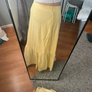 Gul långkjol med volang - Säljer en ljusgul långkjol med bred midja och volang nertill. Kjolen har en mjuk och flowy känsla, perfekt för varma dagar. Enkel och stilren design som är lätt att matcha med olika toppar. Har slits längs ner på båda sidorna, och en liten liten fläck på sidan som inte är så tydlig syns på sista bilden. Storlek L men stretchig så passar nog från s-l!