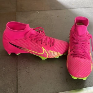 Nike Air zoom mercurial  fotbollsskor i neonrosa - Säljer ett par Nike fotbollsskor i en riktigt snygg neonrosa färg med gula detaljer och snörning. Skorna har högre skaft och är perfekta för dig som vill sticka ut på planen. Swoosh-loggan syns tydligt på sidan. Är använda ca 3månader För spel på naturgräsplaner