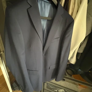 Mörkblå kostym från Dressmann slim fit - Säljer en stilren mörkblå kostym från Dressmann i slim fit-modell. Kavajen har klassisk krage, tre fickor framtill och matchande byxor i storlek 34/30. Perfekt för bal eller fest. Kostymen är helfodrad och har snygg passform. Inköpt våren 24 och har använts 2 gånger. Nypris då 2500:-