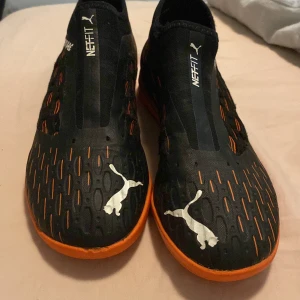 Inomhus fotbollskor/ puma future - Säljer ett par svarta fotbollsskor från Puma med orange sula och detaljer. Skorna har NEFIT-teknologi och är märkta med 'FUTURE' på sidan. De har en låg profil och är designade för inomhusspel.