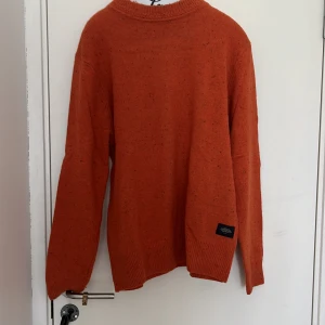 Orange stickad tröja från Scotch & Soda - Säljer en orange stickad tröja från Scotch & Soda. Tröjan har rund halsringning och ribbade muddar vid ärmslut och nederkant. Perfekt för kyligare dagar och enkel att matcha med jeans eller chinos.
