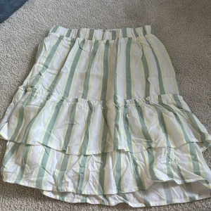 Randig volangkjol med resårmidja - Säljer en söt kjol med volang och resår i midjan. Kjolen har vita, gröna och gula ränder och är i en luftig bomullskvalitet. Perfekt för varma dagar och enkel att matcha med en enkel topp.