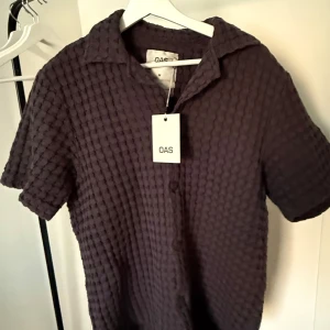 OAS Nearly Black Cuba Waffle shirt - Säljer en mörkgrå (nästan svart) kortärmad pikéskjorta från det exklusiva svenska märket OAS. Skjortan är tillverkad i 100% våfflad bomull, vilket ger en unik och härlig textur som sticker ut. Med klassisk krage och knappar framtill passar den perfekt för en dag vid stranden eller pool cluben.  Nypris: 1400 kr Skjortan är helt oanvänd med tags. Perfekt för dig som värdesätter kvalitet, komfort och en cool, minimalistisk stil!   Skicka DM vid intresse eller frågor. Först till kvarn! 🪭🪩✨
