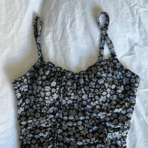 Blommig svart klänning med smala axelband - Säljer en svart klänning med små blommor i vitt, blått och grönt. Klänningen har smala axelband, rynkade detaljer och en figurnära men stretchig passform. Perfekt för sommarens varma dagar!