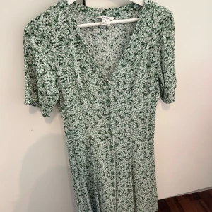 Grön blommig klänning från Monki - Söt grön klänning från Monki med små vita blommor, v-ringning och knappar framtill. Klänningen har korta ärmar och en lätt utställd passform som ger en avslappnad och somrig känsla. Storlek XS passar även S. Aldrig använd. 