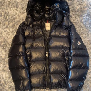 Svart pufferjacka från Moncler - Säljer en svart pufferjacka från Moncler med glansig finish och huva. Jackan har dragkedja framtill och två sidofickor med dragkedja. Klassisk Moncler-logga på ärmen. Perfekt för kyliga dagar.