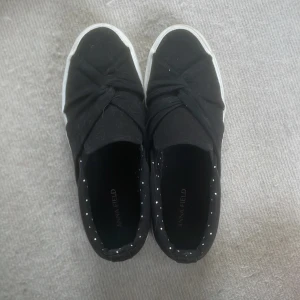 Svarta slip-on sneakers från Anna Field - Svarta slip-on sneakers från Anna Field med knutdetalj framtill och vit sula. Insidan har ett prickigt mönster i vitt. Skorna har rund tå och platt sula, perfekta för en avslappnad stil. Knappt använde, nypris är 339kr, pris kan diskuteras.