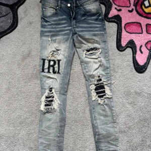 Blå slitna jeans från Amiri med broderad text - Storlek 28/XS. Säljer ett par blå jeans från Amiri med kraftiga slitningar, hål och broderad text 'IRI' på vänster ben. Jeansen har skinny fit och klassisk femficksdesign. Perfekt för dig som gillar en edgy streetstil.