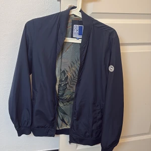 Mörkblå bomberjacka från Jack & Jones - Säljer en stilren mörkblå bomberjacka från Jack & Jones. Jackan har dragkedja framtill, ribbade muddar vid ärmslut och midja samt en snygg logotyp på ärmen. Perfekt för dig som vill ha en klassisk look med modern touch. Hör gärna av er vid frågor eller funderingar!😁