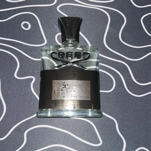 Creed Aventus Eau de Parfum - Lyxig parfymflaska från Creed, modellen Aventus. Flaskan är genomskinlig med svart etikett och svart kork. Märkets logga syns tydligt på framsidan och etiketten har en silverdetalj med en ryttare. Perfekt för dig som gillar exklusiva dofter. Utan box eller kvitto