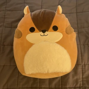 Squishmallow Melzie ekorre - Supermjuk och gosig Squishmallow i form av en ekorre som heter Melzie. Den har bruna och beige färger, stora svarta ögon, rund mage och en fluffig svans. Perfekt som kramdjur eller dekoration i sängen.