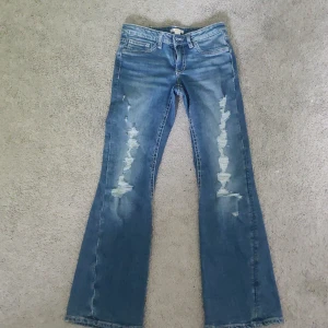 Blå bootcut jeans från Gina Tricot - Säljer ett par blå bootcut jeans från Gina Tricot med slitna detaljer framtill och bakfickor med dital och knappar. Klassisk femficksmodell och normal passform.  stolek 164