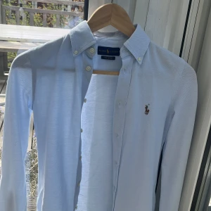 Ljusblå skjorta från Ralph Lauren - Klassisk ljusblå skjorta från Ralph Lauren med button-down krage och broderad logga på bröstet. Skjortan är långärmad och har knappar framtill. Perfekt för en stilren och tidlös look.