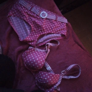 Rosa bikini med vita prickar och randiga detaljer - Säljer en söt rosa bikini med vita prickar och randiga detaljer på både överdel och underdel. Överdelen har vadderade kupor och justerbara axelband, underdelen har en dekorativ randig midjerem med spänne.