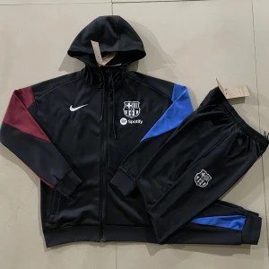 Svart Barcelona hoodie med dragkedja - Säljer en svart hoodie från Nike med FC Barcelona-logga och Spotify-tryck på bröstet. Tröjan har dragkedja, huva och färgdetaljer i vinrött och blått på ärmarna. Perfekt för fotbollsfans som vill ha en sportig look.