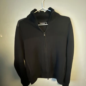 Svart cardigan från J.Lindeberg - Svart cardigan från J.Lindeberg. Tröjan har dragkedja framtill och är långärmad. Snygg och stilren design med diskreta axelpartier i annat material.