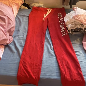 Röda mjukisbyxor från Abercrombie & Fitch - Röda mjukisbyxor från Abercrombie & Fitch med vit dragsko i midjan och tryckt logga längs benet. Sköna och stretchiga med breda ben och texten EST 1892 bak. Perfekta för chill eller träning.