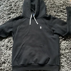 Svart hoodie från Polo Ralph Lauren - Säljer en svart hoodie från Polo Ralph Lauren med klassisk vit broderad logga på bröstet. Tröjan har huva med vita snören och en stor magficka framtill. Perfekt för en avslappnad och stilren look. 