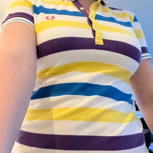 Randig piké t-shirt från Fred Perry - Säljer en färgglad piké t-shirt från Fred Perry med breda ränder i gult, blått, lila och vitt. Klassisk krage med knappar och broderad logga på bröstet. Perfekt för dig som gillar sportig och färgstark stil! Funkar för alla kön. 