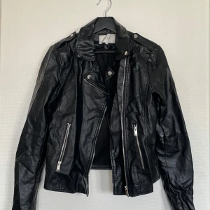 Svart skinnjacka från VILA - Jackan har sned dragkedja, krage med knappar och två fickor med dragkedjor framtill. Snygga detaljer på axlarna och smal passform.  Använd väldigt sparsamt.