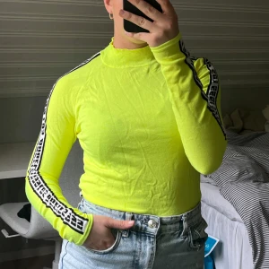 Neongul långärmad topp med tryck på ärmarna - Neongul långärmad topp med hög hals och svarta/vita tryckta band längs ärmarna. Köpt på HM med aldrig använd, utan har legat i garderoben. Därav att den är skrynklig💛