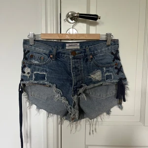 Blå jeansshorts med snörning från One Teaspoon - Säljer ett par blå jeansshorts från One Teaspoon med råa kanter, slitningar och cool snörningsdetalj i sidorna. Shortsen har fem fickor och låg midja. Perfekta för en avslappnad och edgy stil. Säljer för att de är för stora!