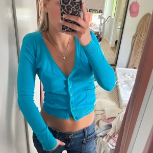 Turkos croppad kofta - Säljer en turkos croppad kofta med v-ringning och långa ärmar. Koftan har knappar framtill och är i ett mjukt, stretchigt material. Perfekt att styla med jeans eller kjol för en fräsch look.