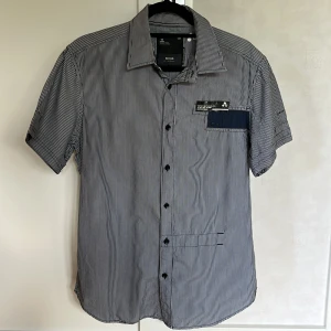 Vintage G-Star skjorta - Säljer en snygg kortärmad skjorta från G-Star i svartvitt randigt mönster. Skjortan har knappar framtill och en bröstficka med detalj. Tillverkad i 100% bomull, perfekt för varmare dagar.