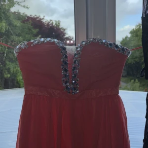 Röd axelbandslös långklänning med paljettdetaljer från NLY Eve - Säljer en röd axelbandslös långklänning från NLY Eve med glittriga paljettdetaljer längs urringningen. Klänningen har dragkedja på ryggen och är tillverkad i ett glansigt material som ger en lyxig känsla.